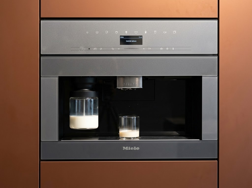 Miele Einbau-Kaffeevollautomat mit Milchbehälter in einer Ausstellungsküche von Kitchen Concept, umgeben von kupferfarbenen Fronten