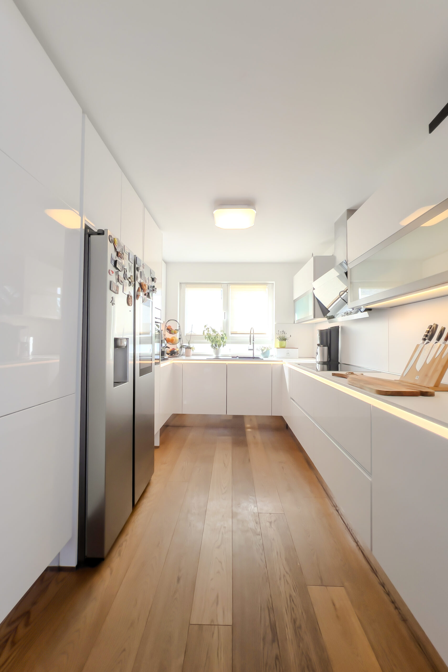 Helle moderne nobilia U-Küche in Weiß Hochglanz mit warmweißer LED-Griffmuldenbeleuchtung, Side-by-Side-Kühlschrank und Holzboden.